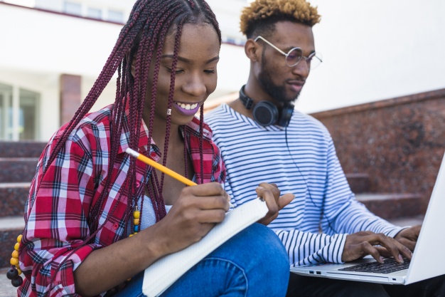 Uma moça negra escreve num caderno e um rapaz negro digita no laptop