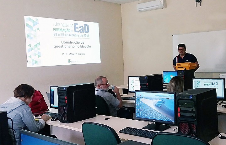 Professor à frente de uma sala cheia de computadores, com uma apresentação de slides ao fundo