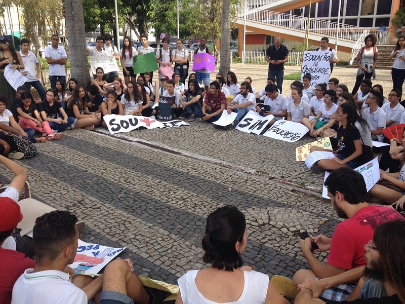 Alunos sentados ao chão e outros em pé, em roda, com cartazes contra medidas do governo